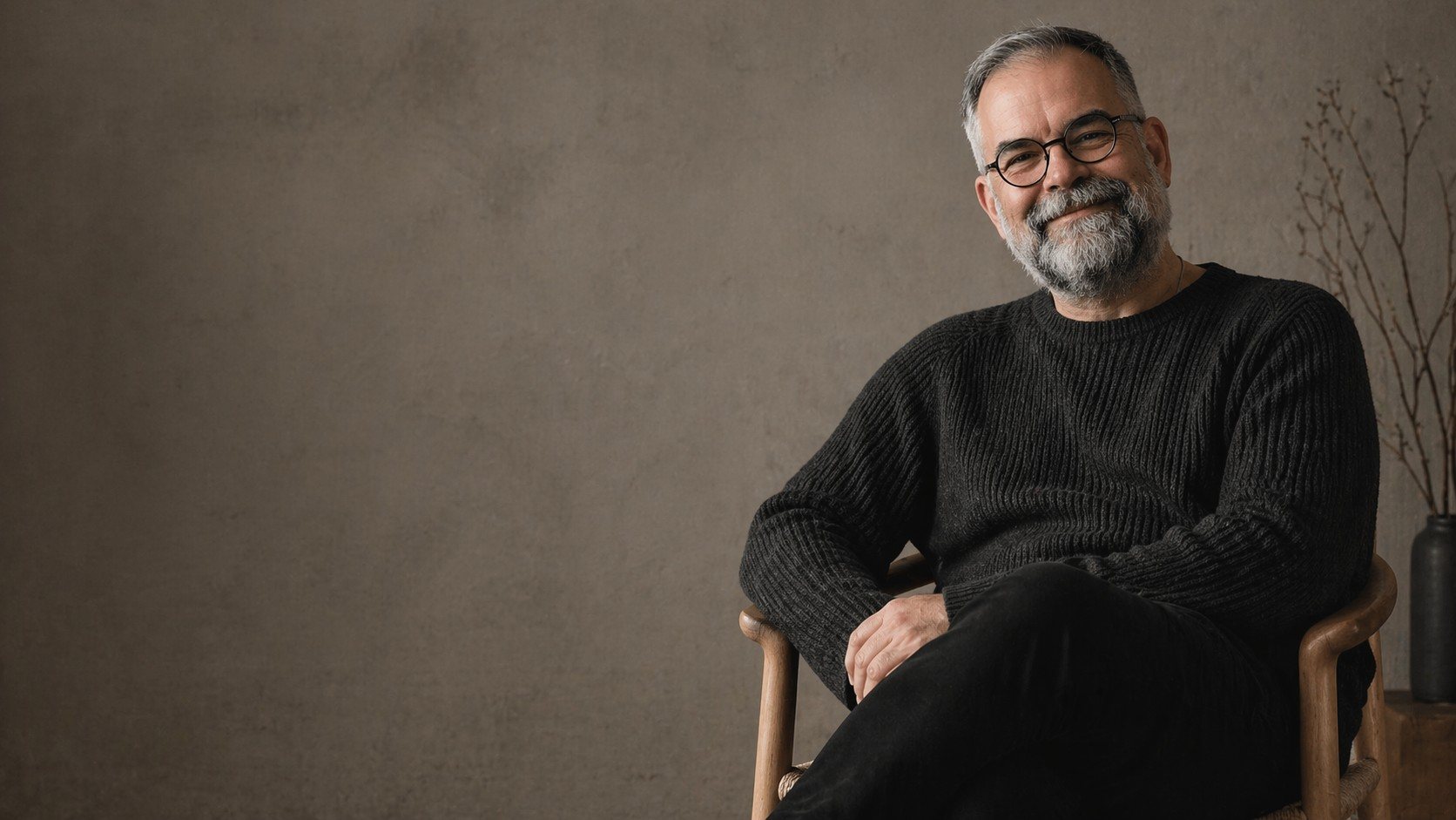 Ericson seated in a wooden chair, wearing a black sweater and round glasses, smiling